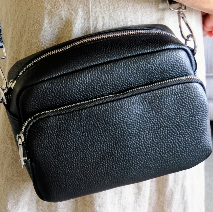 A black pebbled leather crossbody bag with silver zippers and hardware, shown worn across a person's torso in a beige dress.