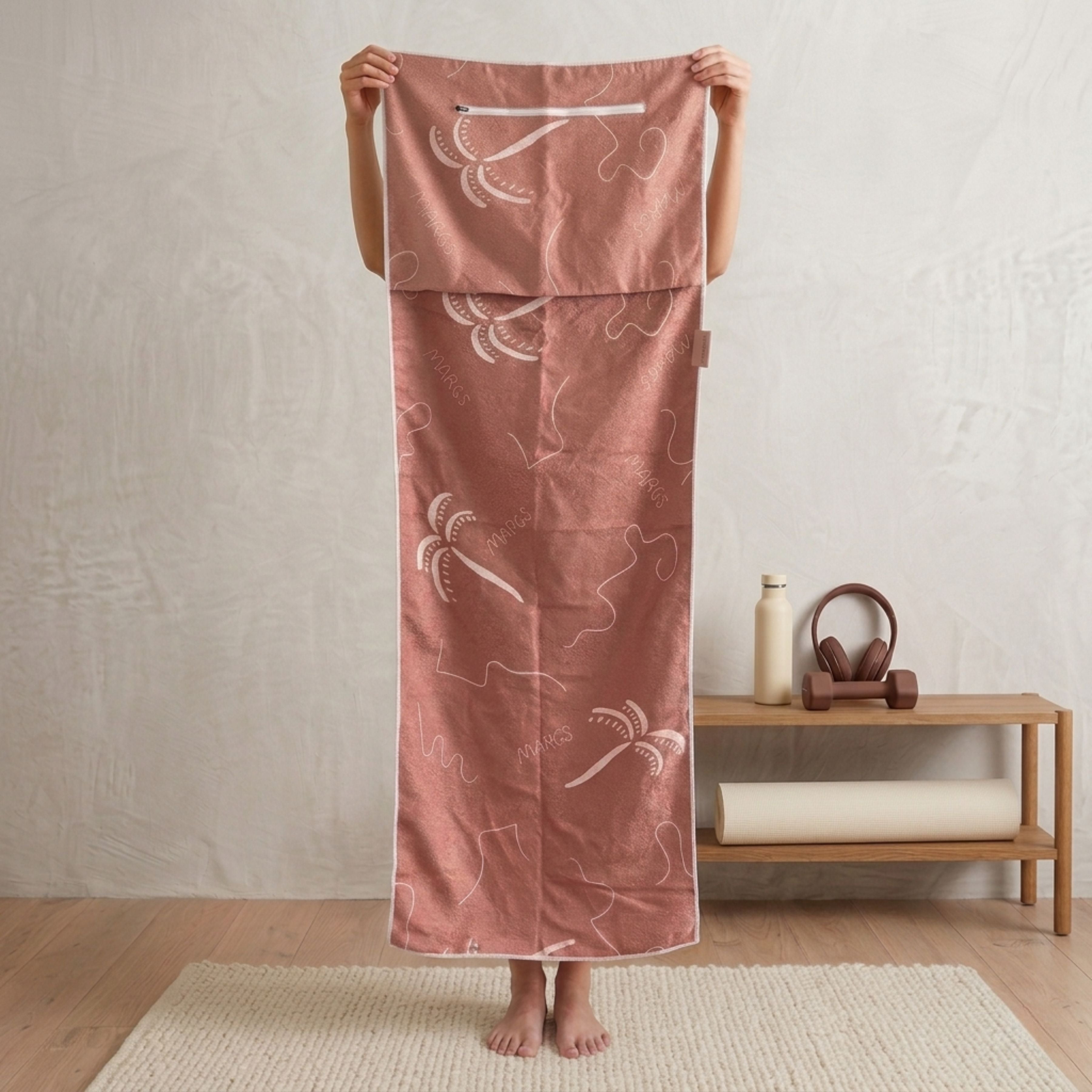 A minimalist wellness or yoga space with a person holding a microfibre gym towel. In the background, there is a small wooden shelf holding pink headphones, a pink dumbbell, a white water bottle, and a rolled yoga mat labeled “ZEN WAVE.” The floor has a natural woven rug, and the neutral wall creates a calm, studio-like atmosphere.