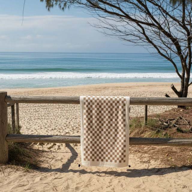 The cream and brown tones blend seamlessly with the surrounding sand, making the towel feel integrated into the beach environment rather than visually separate from it.