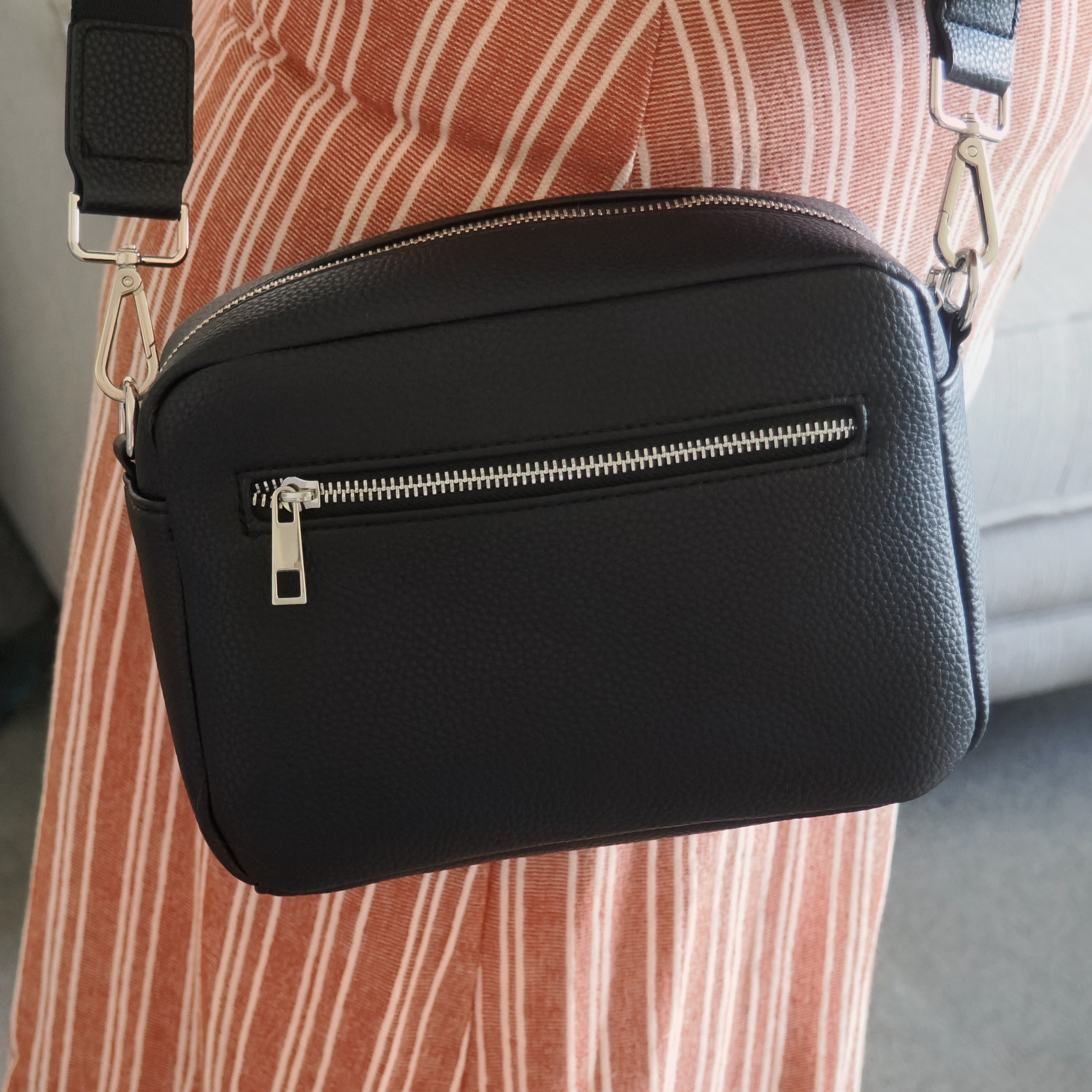 A black pebbled faux-leather crossbody bag with a silver zipper and hardware, worn across a person's torso over a rust-orange and white striped dress.