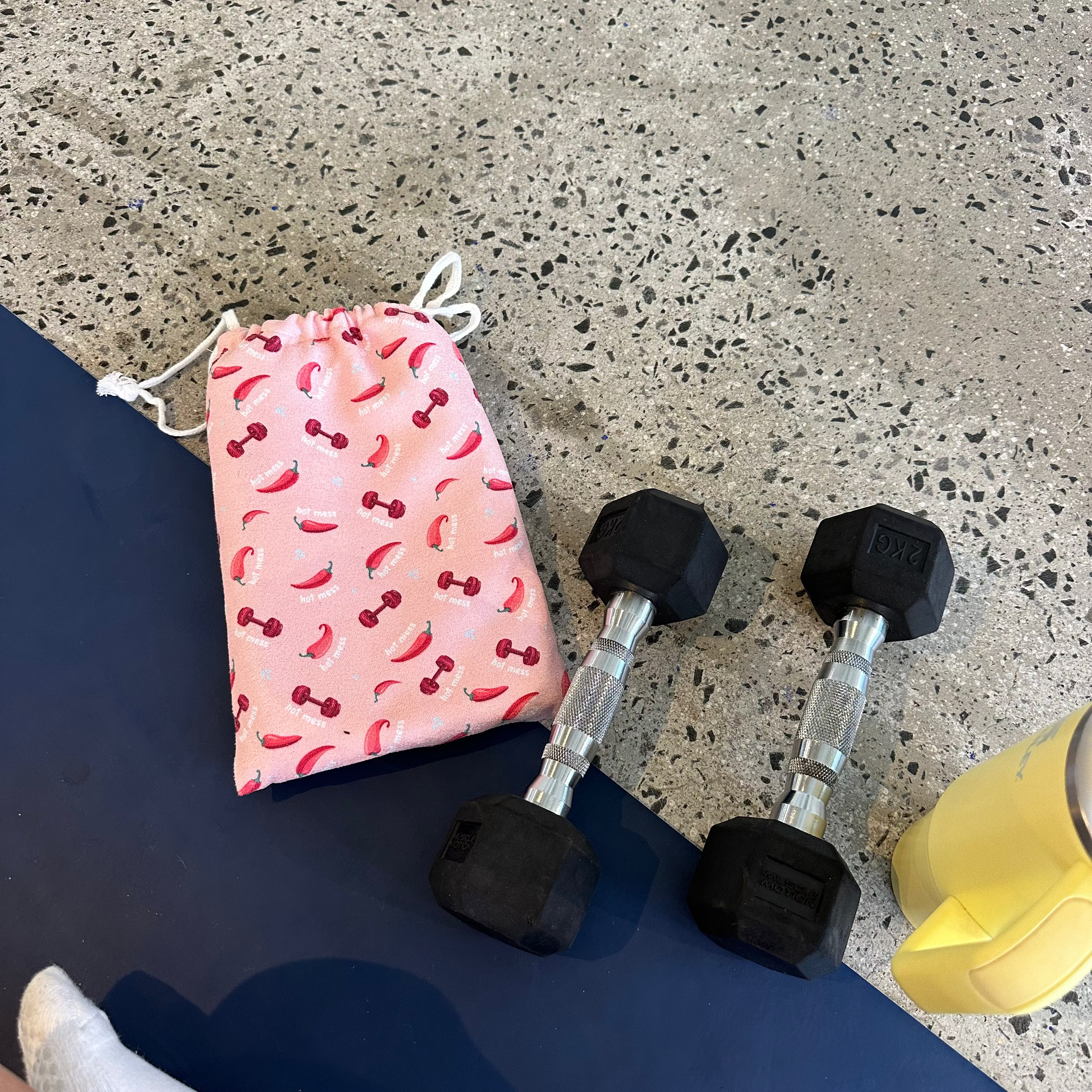Two black hex dumbbells with silver metal handles are lying on the floor with a yellow Stanley tumbler and a cute pink gym towel bag.