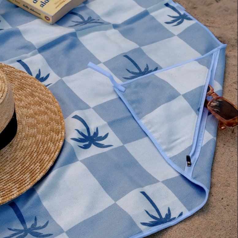 Blue checkered picnic blanket with a straw hat, book, and glass on a sandy surface